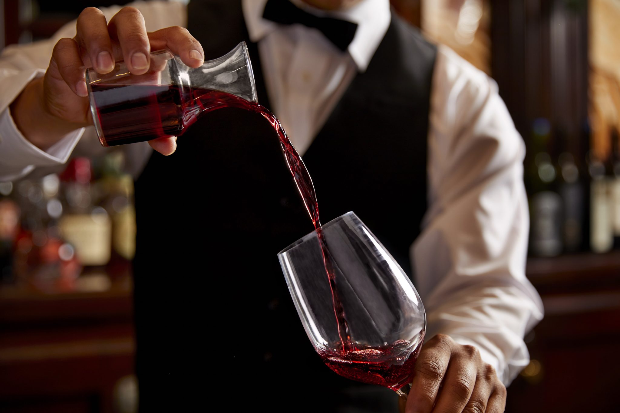 A bartender wearing a vest and bowtie pouring red wine from a carafe into a glass