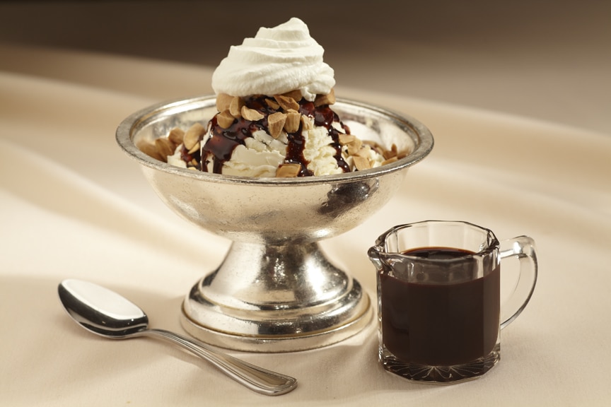 A chocolate sundae in a silver bowl topped with whipped cream and nuts, with a side of hot fudge.