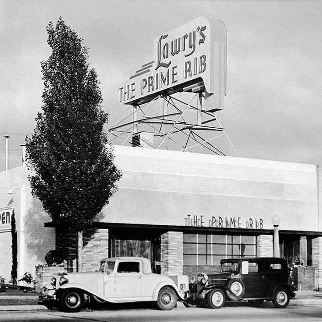 Vintage photo of the original Lawry's The Prime Rib building with two antique cars in front.