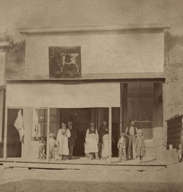 Vintage photo of a storefront with several people standing on the wooden sidewalk in front.