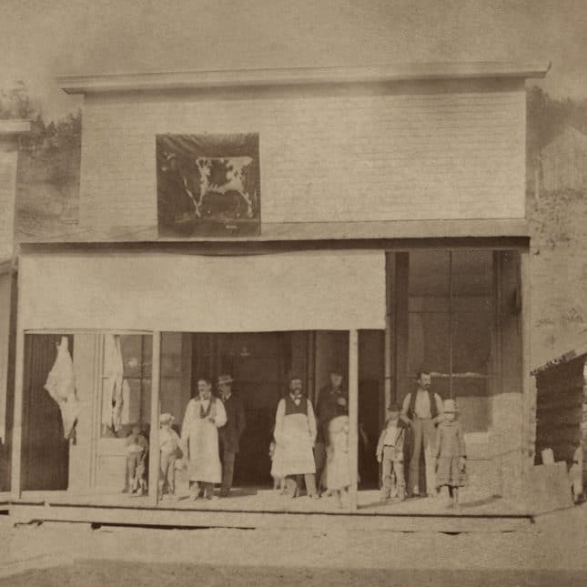 Vintage photo of a storefront with several people standing on the wooden sidewalk in front.