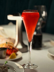 A vibrant, ruby-red sparkling cocktail served in a sleek, modern flute, part of a Wizard of Oz-inspired collection at Lawry's The Prime Rib Las Vegas. The drink features a subtle shimmer and is set against a backdrop of a gourmet "surf and turf" dinner including lobster tail, lemon, and prime rib.