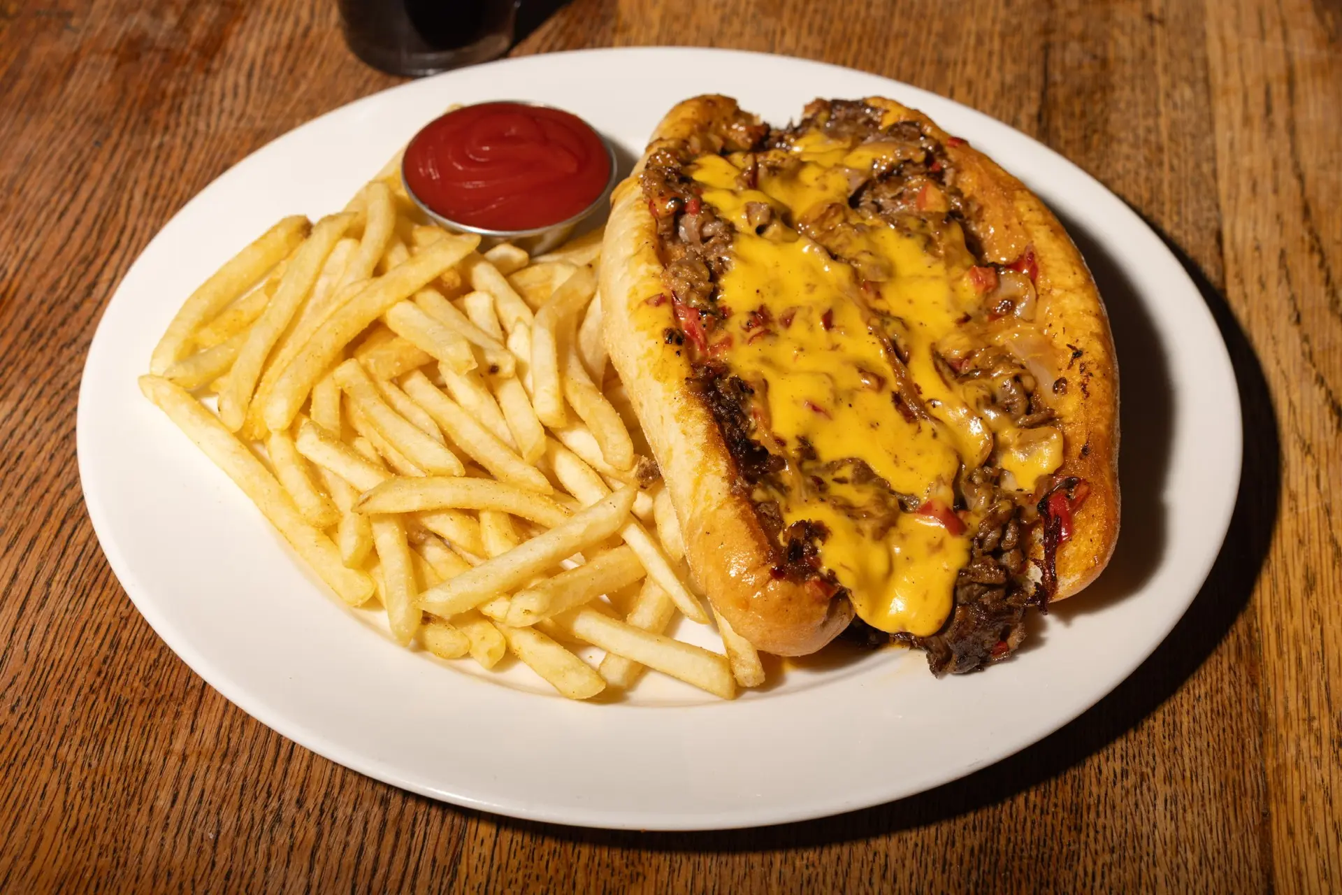 Prime Rib Philly cheesesteak sandwich smothered in melted cheese sauce with sautéed peppers and onions, served with crispy french fries and ketchup on a white plate